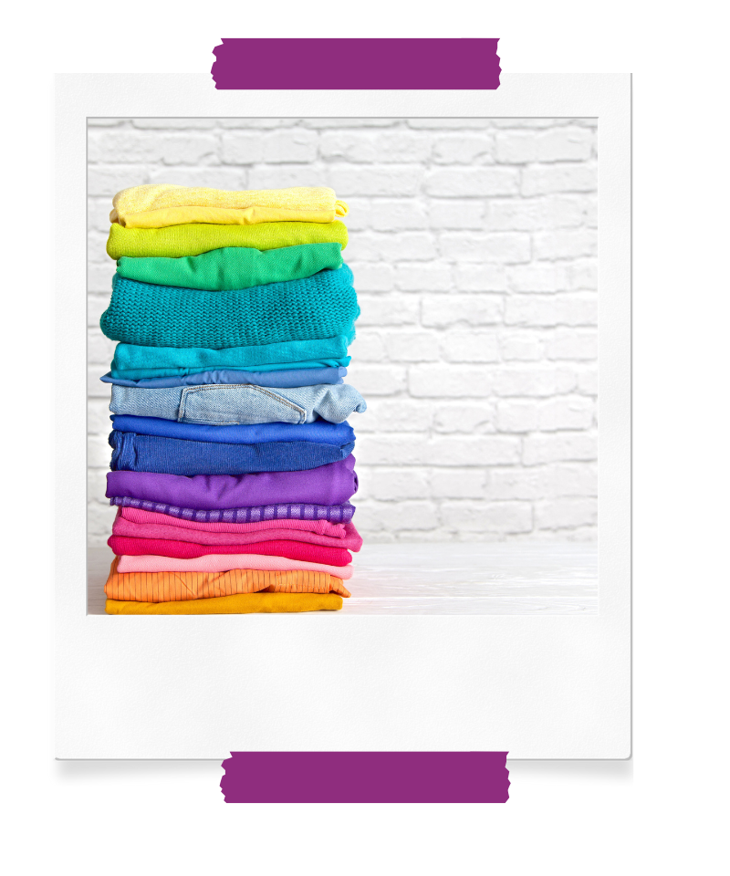 Ein Stapel leuchtend bunter Kleidung, ordentlich gestapelt auf einem hellen Tisch vor einer weißen Backsteinwand. Die Farben reichen von Gelb über Grün, Blau, Lila bis hin zu Pink und Orange.