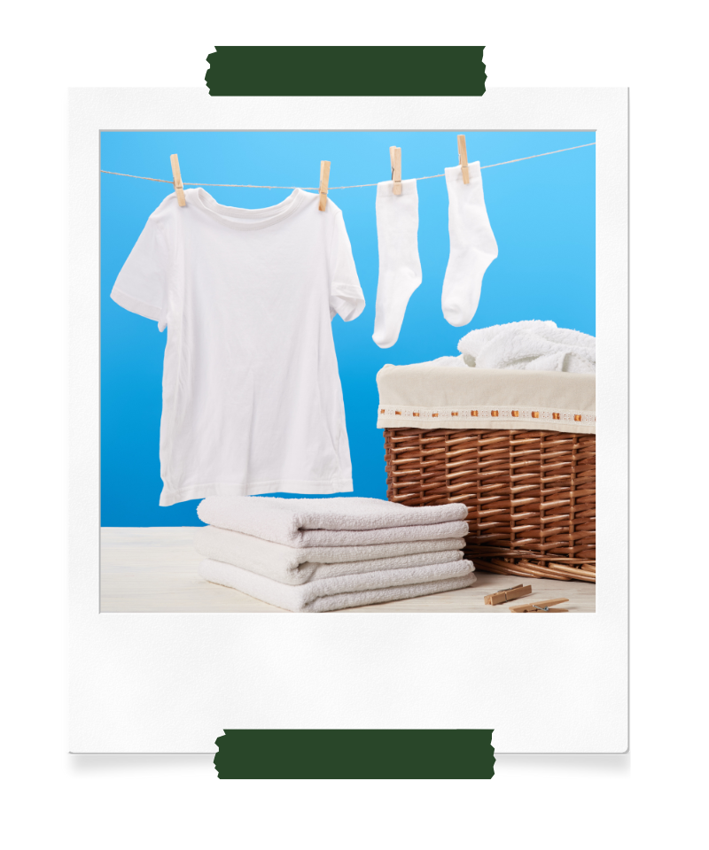 Frisch gewaschene weiße Wäsche, darunter ein T-Shirt und Socken, hängen auf einer Wäscheleine vor einem blauen Hintergrund. Daneben liegt ein Stapel weißer Handtücher und ein geflochtener Wäschekorb auf einem Tisch.